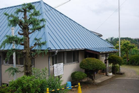 青い屋根にベージュの壁の建物の周りにたくさんの木が植えられており、玄関の入口にも屋根が付いている飯山中部児童館の外観写真