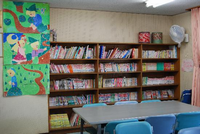 壁にはちぎり絵の作品がが飾られ、沢山の本が並んでいる本棚が設置されており、本棚の前にテーブルと椅子が置かれている館内（図書室）の写真