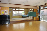 広い遊戯室に黒板とピアノが置かれたおり、その奥に畳に部屋がある中戸田児童館の遊戯室の写真