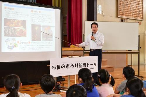 市長が体育館に集まった生徒たちの前で指し棒を使ってプロジェクタースクリーンを指しながら話をしている写真