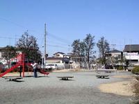 住宅街の一角にある樹木が点在している園内右側に2つの背もたれのあるベンチ、中央に3つの背もたれのないベンチが並んでおり、奥にブランコ、左側手前に赤色のすべり台が設置されている、しもこが公園の写真