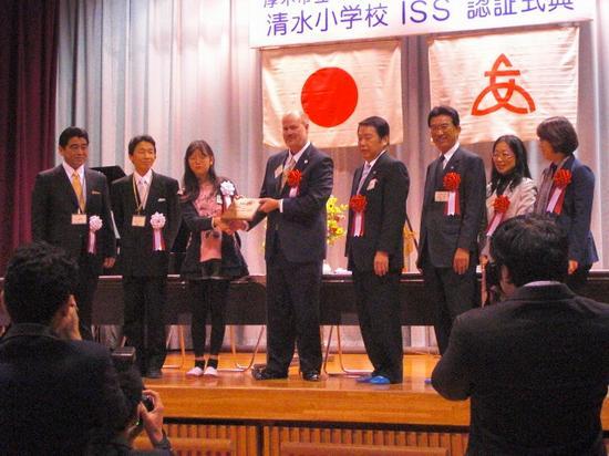 壇上に授与式の関係者が並んでいます。中央の男女が認証盾を持って、胸章を付けた8人で記念写真