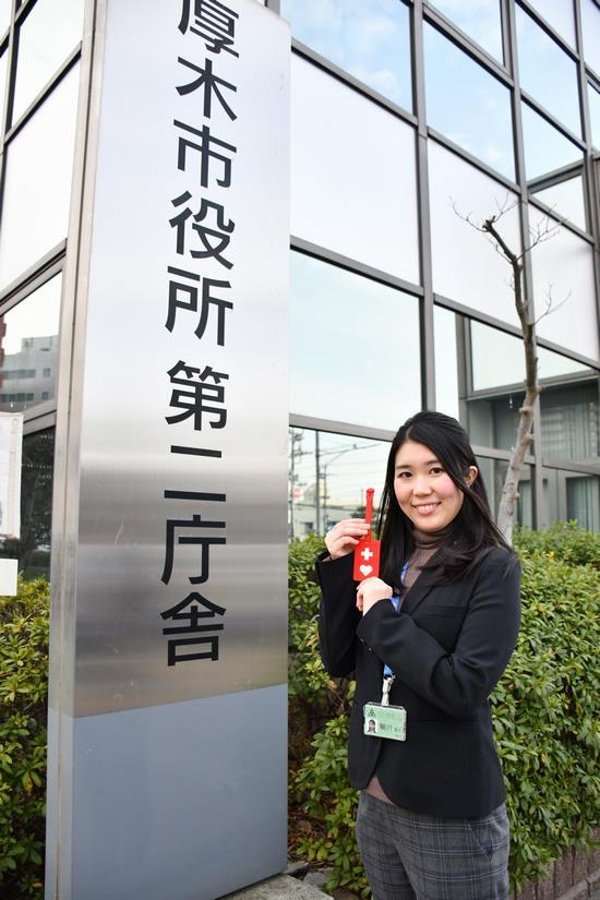 厚木市役所第二庁舎の看板の横にヘルプマークをもって立っている女性職員の写真