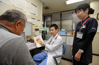 診察室にて男性医師が年配の男性の診察を行っており、女性看護師が傍に立っている写真