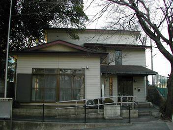 入口に黒い柵がしてある先に手すり付きのスロープと階段が玄関まである白い壁の温水老人憩の家の外観写真