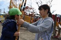 保育園の園庭で緑色の帽子を被った男の子が竹馬に乗っているのを女の先生が支えている写真
