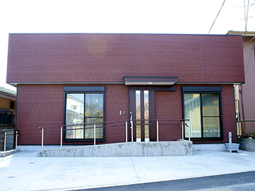 壁がレンガ色で、左右に窓ガラス中央に玄関があり、建物の前に手すり付きのスロープ設置されている、関口フェルマータ小規模保育園の建物外観の写真