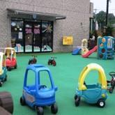 人工芝の広がる園庭に並べられた色とりどりの車の園児が乗れる車の4輪車やすべり台などの写真