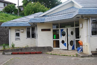 白い壁に青い屋根で、入口のドアに丸い窓がある宮の里児童館正面の写真