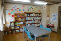 壁には折り紙で作られたたくさんの花が貼ってあり、その下に本棚や引き出しが設置されている手前にテーブルと椅子が並べられている妻田児童館の図書室の写真