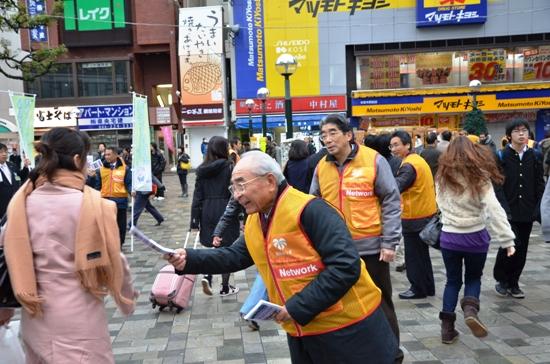 オレンジ色のベストを着た男性たちが街頭で市民に資料を配布している写真