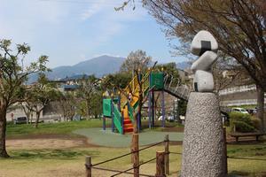 「昇」の作品を撮影した写真。作品は公園の遊具の近くにある仕切りの中に設置されており、丸い形をした灰色の土台の上に薄い灰色のおにぎりを表現したようなオブジェが3つ重なって置かれている。