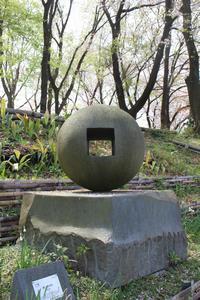 「球＝888」の作品を撮影した写真。作品は公園の木の近くに設置されており、薄い四角の形をした灰色の土台の上に、薄い緑色の真ん中に穴の開いた球体を表現したようなオブジェが置かれている。