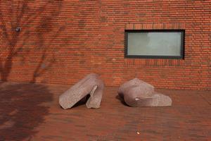 「時間軸－Ａ－3」の作品を撮影した写真。作品は文化会館の建物の近くに設置されており、薄い赤茶色の強い重力で四角い石が折れ曲がっていることを表現したようなオブジェが置かれている。