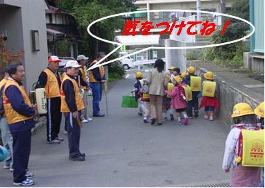 ランドセルを背負い、黄色の帽子を被った子供たちが地域の方々に見守られながら歩いている様子の写真