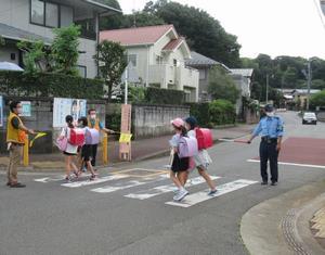 小学生が横断歩道を渡っているのを、警察官や地域の方が黄色の旗を持って見守っている写真