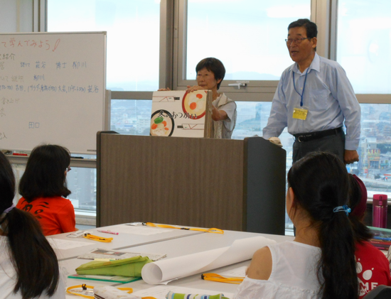 演壇で紙芝居を上演している神奈川県環境学習リーダー会の男性と女性、紙芝居を見ているジュニアエコリーダーの子供たちの写真