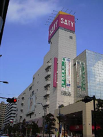 ピンク色の地に文字が白色のSATYのロゴマークの看板が建物の頂上で目立つ大型商業施設やビルが建ち並ぶ市街地の写真