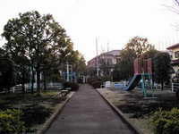 住宅街に囲まれて周囲を樹木が点在している園内中央に通路があります。通路の左側奥にジャングルジム、手前に2つのベンチ、右側にすべり台が設置されているこまつ下公園の写真