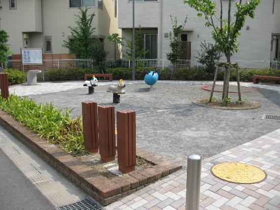 園内左側から水飲み場、2つのベンチ、中央にパンダとコアラのスプリング遊具が設置されている公園を入口から撮影した写真