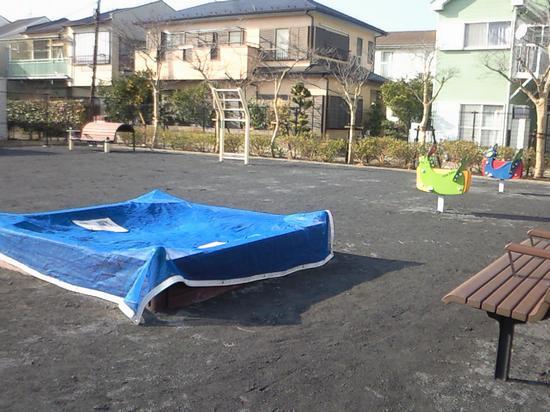 園内右奥に2つのスプリング遊具、手前に青色のカバーが掛けられた砂場、右側にベンチが設置されている、でんえん公園の写真