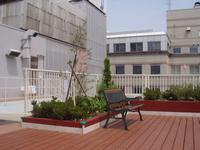 建物の屋上バルコニーに造られたえんじ色のL字型の花壇に、植木や樹木が植えてある写真