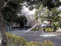 周囲を樹木で囲まれている園内にはベンチが奥に2つ右側に1つあり、中央にすべり台やブランコなどの遊具が設置されている、かやま公園の写真