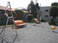 奥にオレンジ色の背もたれが丸くなっている2つのベンチが間を開けて設置されており、ベンチの間と左側に健康遊具、右側に黄色のスプリング遊具がが設置されている園内の写真