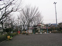大きな樹木や垣根が植えられている園内左側にベンチがあり、奥には横長の東屋が設置されている広場のある戸室しみず公園の写真
