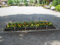 周囲に垣根や樹木が点在している園内左側に2つのベンチが設置され、手前に黄色やオレンジ色、紫色の色とりどりの花が咲いている花壇の写真