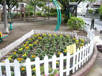 園内に設置された遊具の手前通路沿いの低い白色の枠内に、オレンジ色や黄色などの色とりどりの花が咲いている花壇の写真