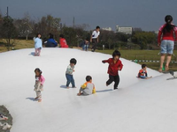 公園のこども広場に設置された雪山の様な形の「ふわふわドーム」の上に、子ども達が飛び跳ねたりして遊んでいる写真