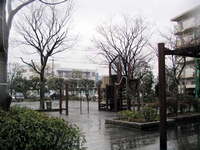 集合住宅の裏手にあり、周囲や園内に垣根や樹木が点在しており、中央には木製の遊具が設置されている岡田南公園の写真