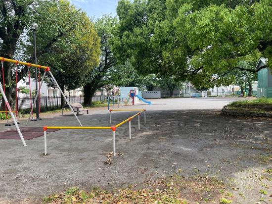 周囲や園内中央に大きな樹木が生えており、道路側の奥にすべり台、手前にぶらんこなどの遊具が設置されている中村公園の写真