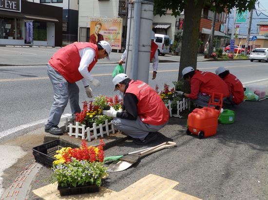 赤いベストを着た男性方が、通りにある街路樹の周りに赤や黄色、白色の花を植えている写真