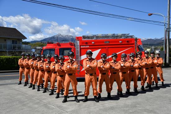 1台の消防自動車の前に災害時のオレンジ色の服にヘルメットを被った18名の隊員が腕を組んで立っている写真