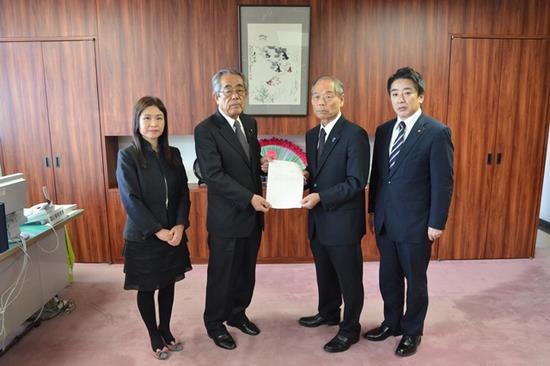 越智一久議長へ答申書を手渡す沼田幸一委員長と右端に黒のスーツ姿の女性と左端のに紺のスーツ姿の男性がたっている写真