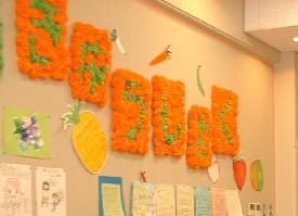 ランチルームの壁にオレンジ色と黄緑色のペーパーフラワーで作った「きゅうしょく」の文字が飾られ、パイナップルなどの食材のイラストも一緒に飾られている写真