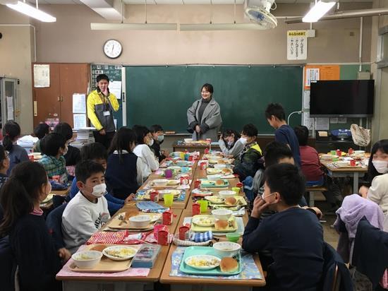 給食がテーブルに並べられている生徒たちの前で先生が生徒に呼びかけをしている横に女性が立っている写真
