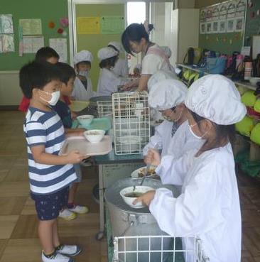 給食着にマスクを着用した給食当番の生徒と並んでお盆におかずをのせていく生徒たちの配膳の写真