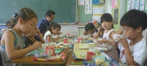 教室で先生と一緒に生徒たちが向かい合って給食を食べている写真