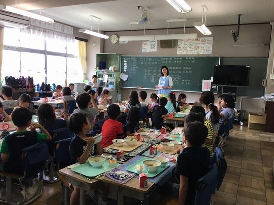 先生と生徒たちの給食の時間に黒板の前で食育の講話をするエプロン姿の栄養士の写真