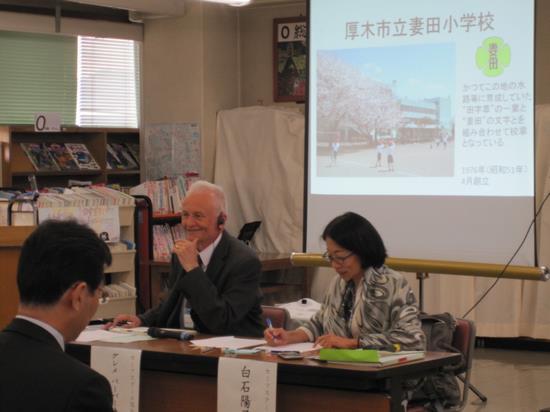 奥のスクリーンに資料が映し出されており、手前に設置した長机の左側に白髪の外国人の男性と、右側に日本人の女性が座って話をしている写真