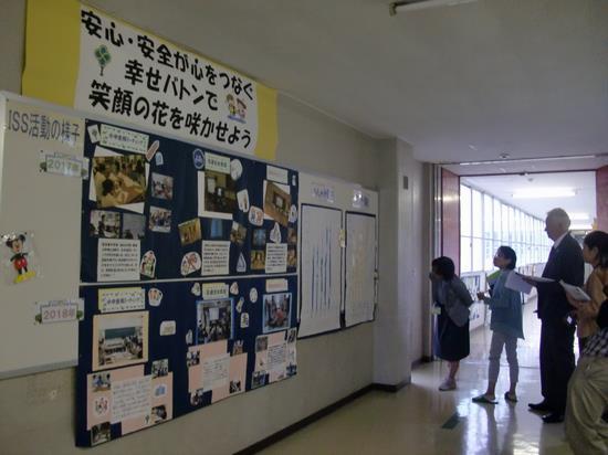 校内の廊下の壁にISS活動の様子を年度別に写真と文字で展示しているのを、審査新の女性と白髪の外国人の男性の審査員が視察している様子の写真