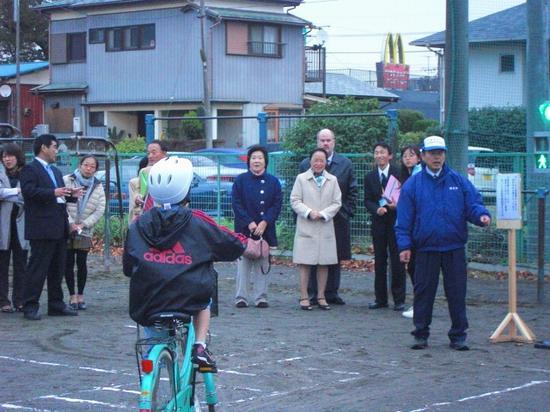 ヘルメットをかぶった小学生が自転車に乗っていて、先生方が見学されている写真