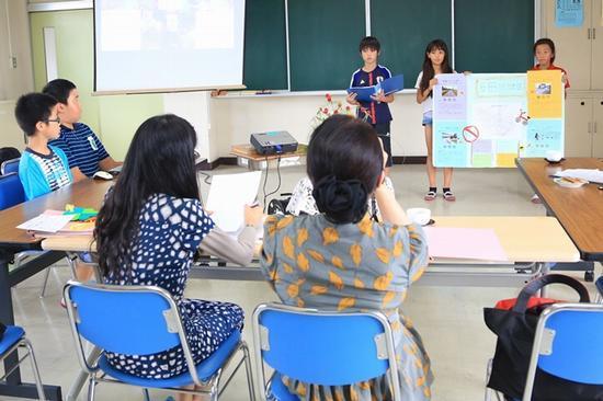 生徒3名が教室の前に立っており、うち2名の女の子が作成した資料を持ち、1名の男の子が青いファイルを持っている、その様子を女性2名と男子生徒2名が見ている写真