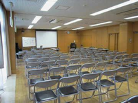 前方にプロジェクタースクリーンが設置され、部屋いっぱいにパイプ椅子が並べられてある集会室の写真