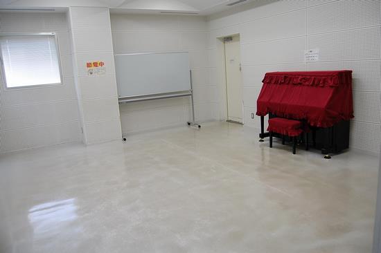 壁際に赤いカバーのかけられたピアノとホワイトボードが設置されており、白色を基調とした会議室C(音楽室2)の写真