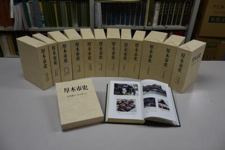 厚木市史民俗編生活記録集13巻中1巻をケースから出して開いている状態と、他12巻は後ろで扇形に並べて立てている写真