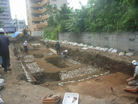 石などが埋まっている建物跡で6名の方が発掘作業をしている写真